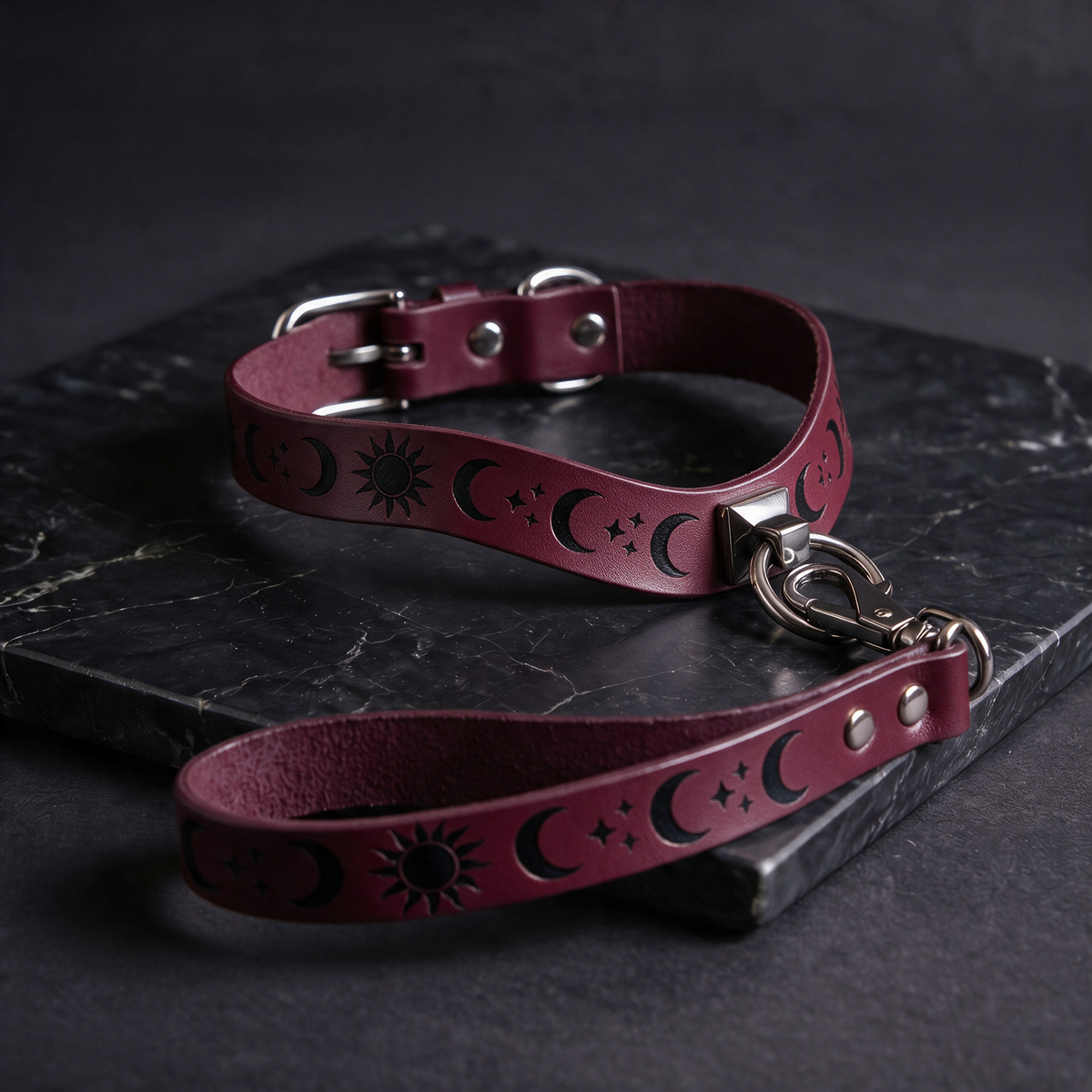 A burgundy leather dog collar with a sun and moon motif, featuring a metal buckle and a matching metal tag, is displayed on a dark surface.