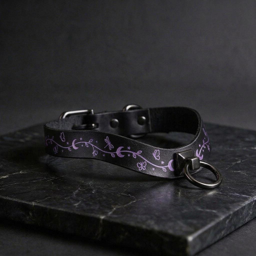 A black leather collar with a silver metal ring and a floral design on it, resting on a dark surface.
