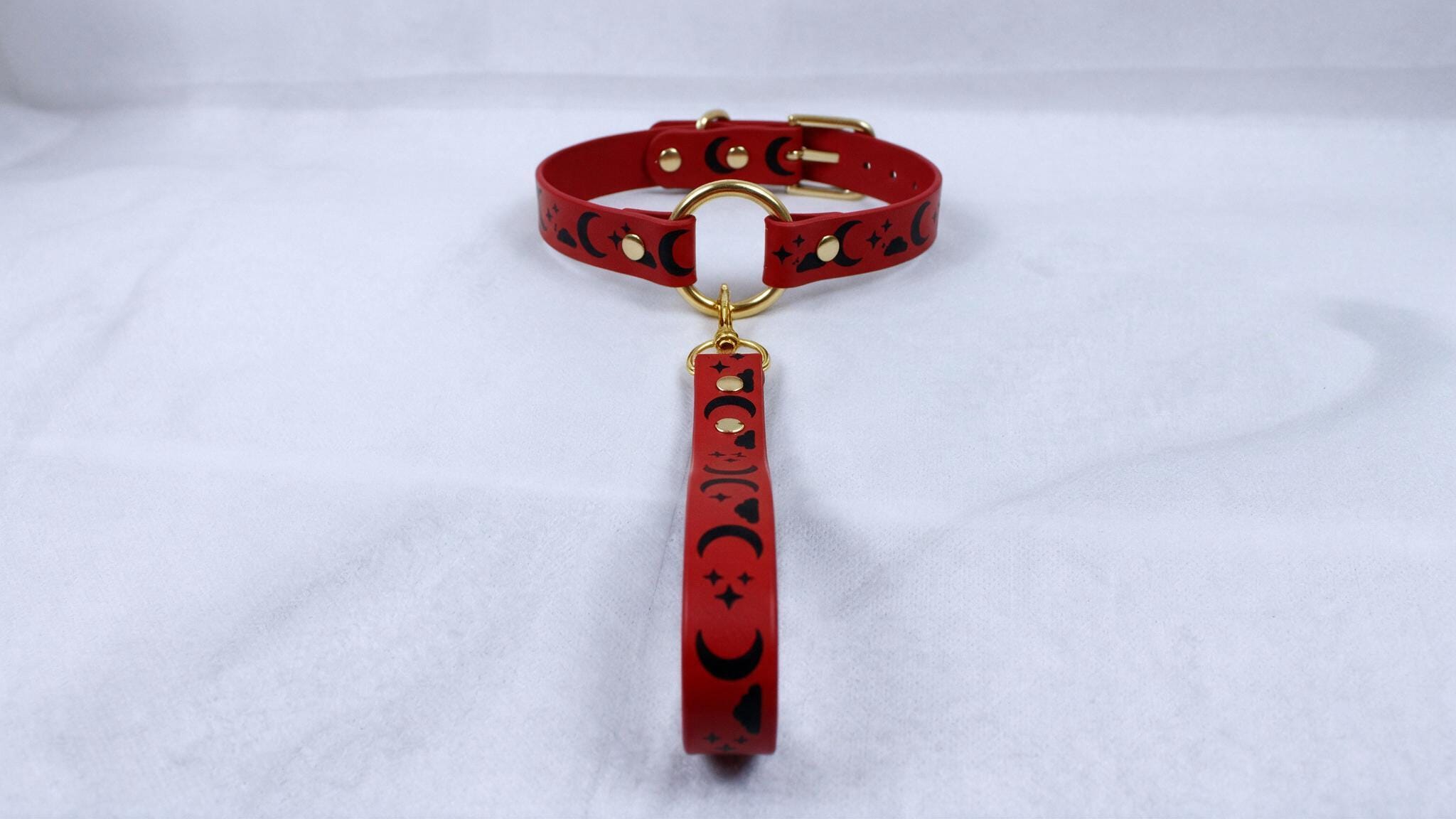 A red collar with a gold-colored buckle and a red strap with black designs is displayed on a white surface.