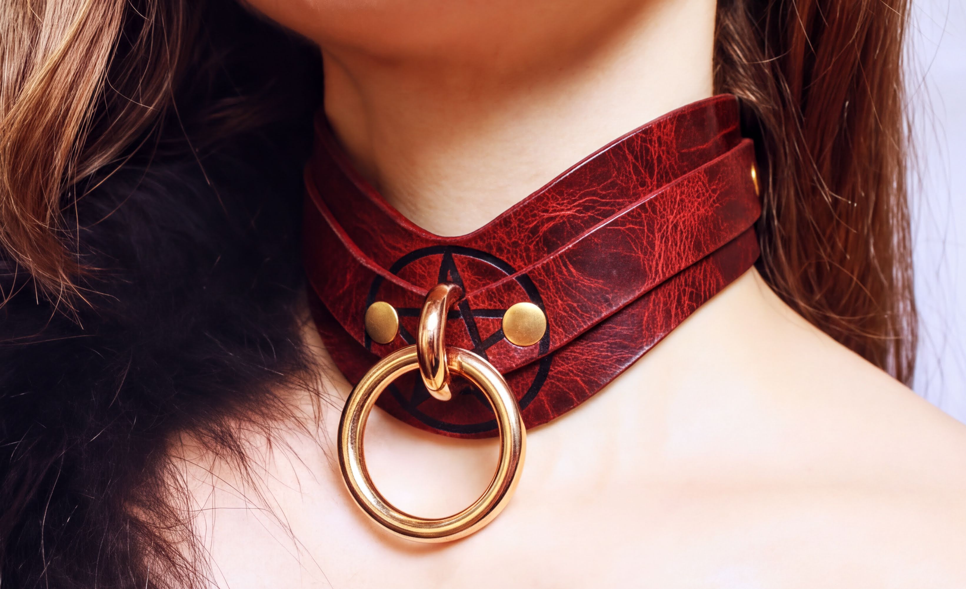 A close-up of a woman's neck wearing a red leather choker necklace with a gold ring and a pentagram symbol.