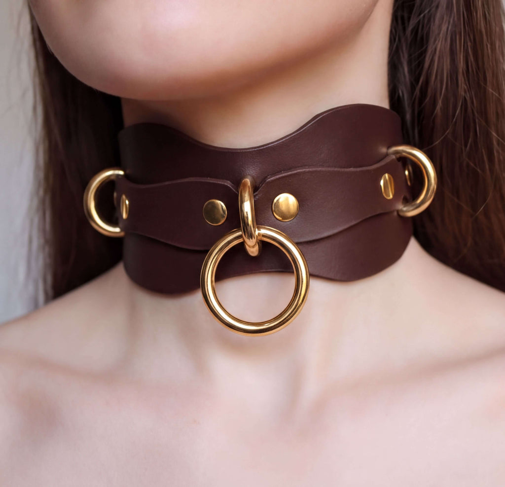 A close-up of a woman's neck wearing a brown leather choker necklace with gold rings and a large circular ring.