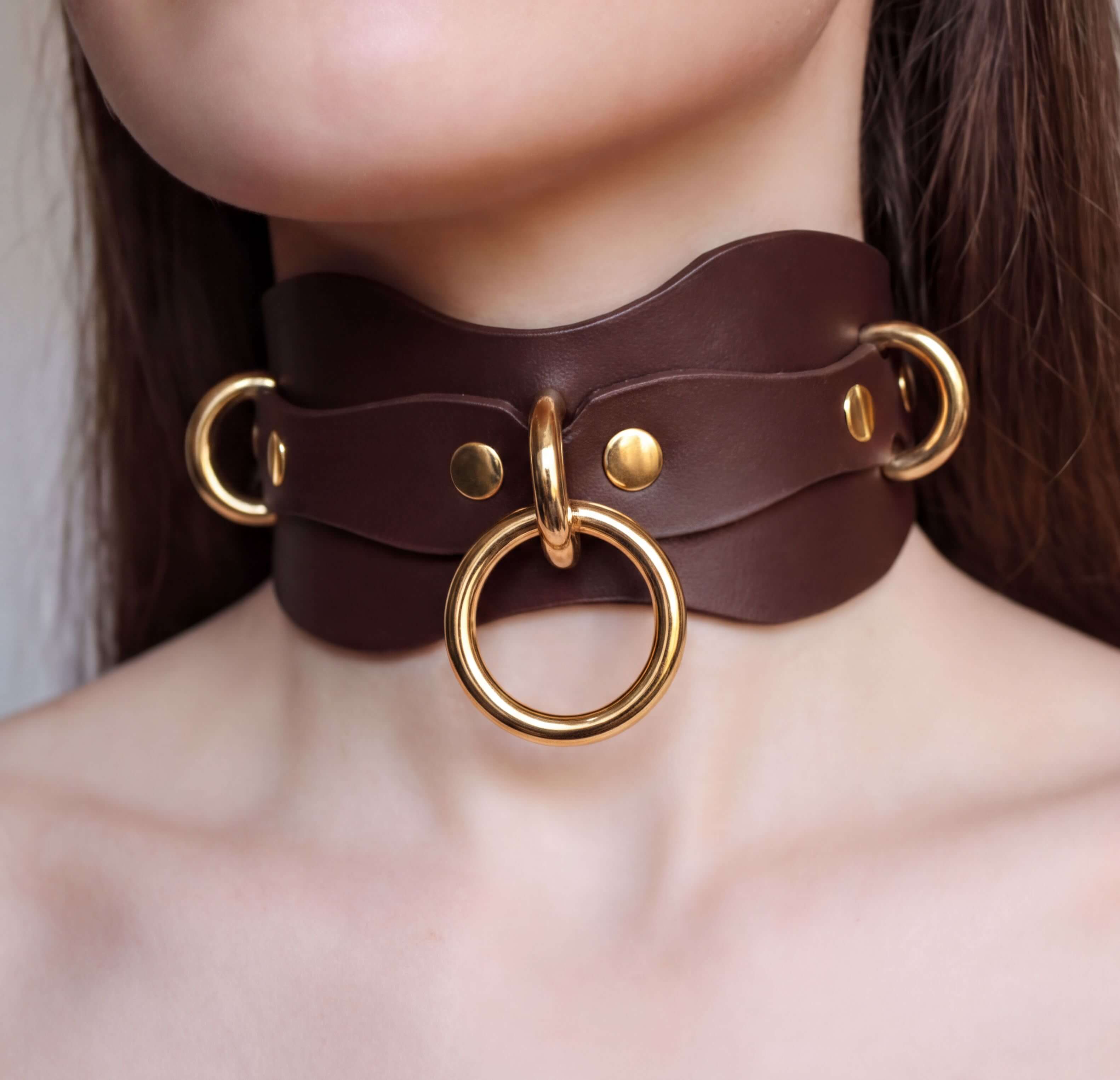 A close-up of a woman's neck wearing a brown leather choker necklace with gold rings and a large circular ring.