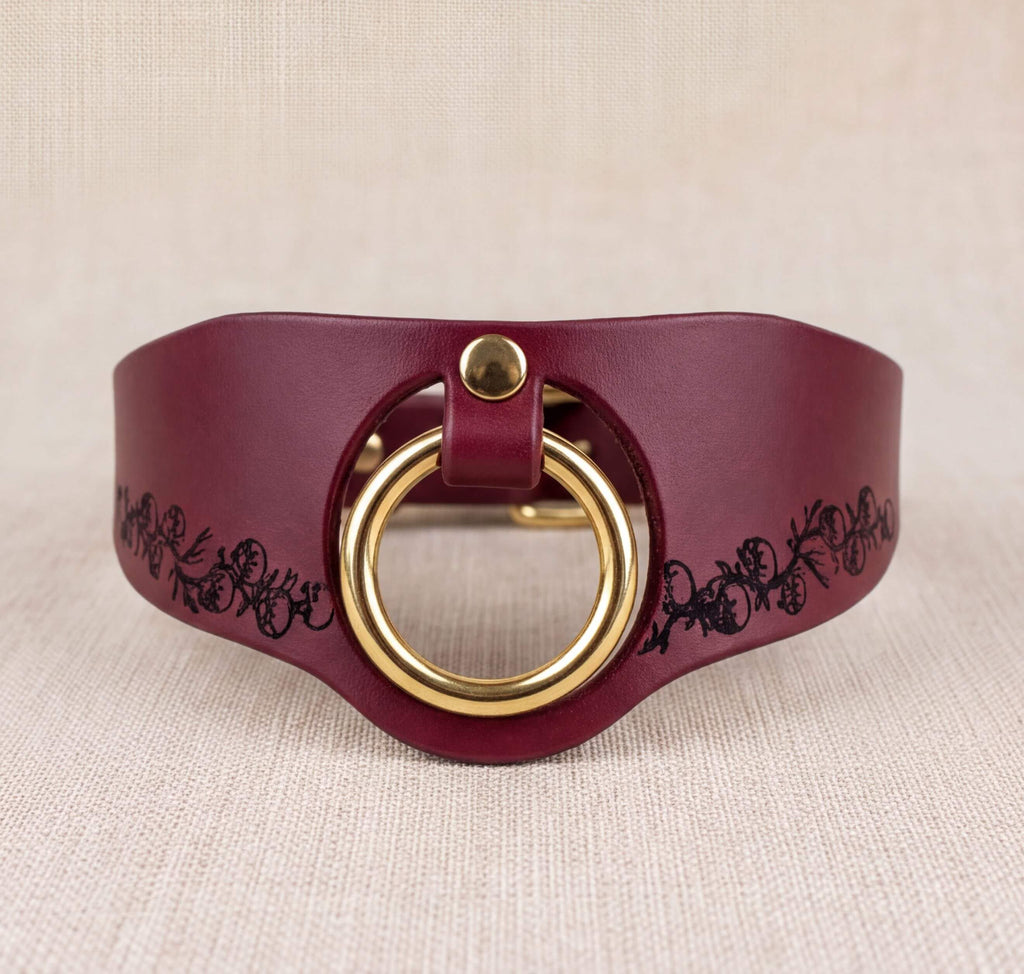 A burgundy leather collar with a gold-colored circular buckle and a floral design on the side.