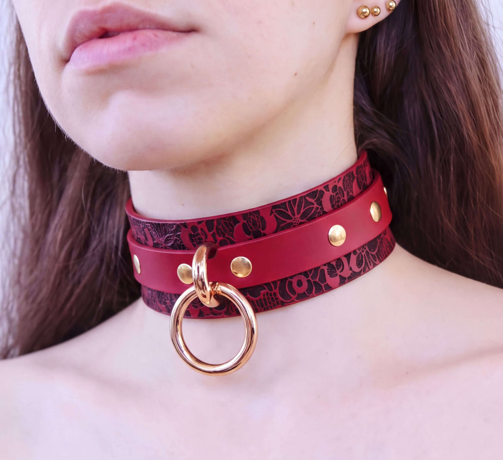 A close-up of a woman's neck and chest area wearing a red choker necklace with a gold ring and studs.