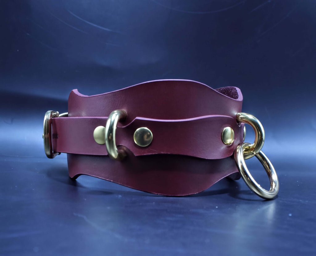 A burgundy leather choker necklace with gold-colored metal rings and buckles is displayed against a dark blue background.