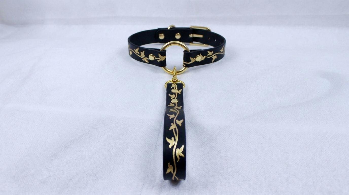 A black collar with gold leaf designs and a gold-colored loop hanging from it.