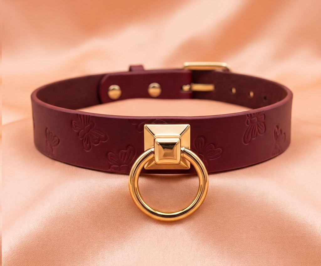 A burgundy leather choker collar with a gold-colored metal ring and a small square cutout in the center.
