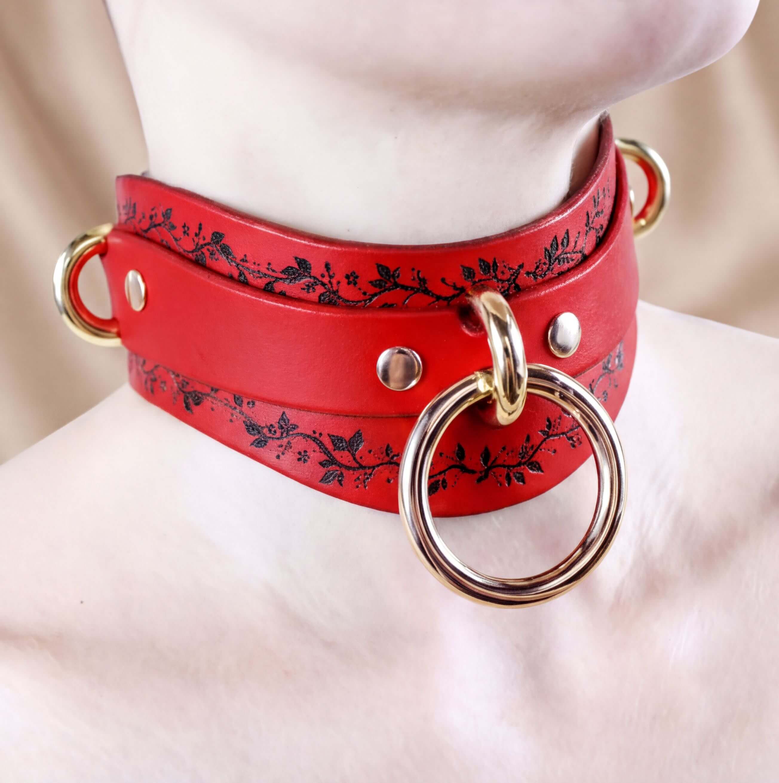 A close-up of a red leather choker necklace with a gold ring and a floral design on the collar.