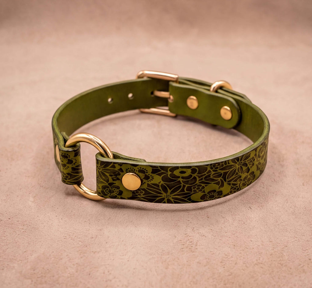 A green leather collar with a floral pattern and gold hardware, including a buckle and two rings, is displayed on a beige background.