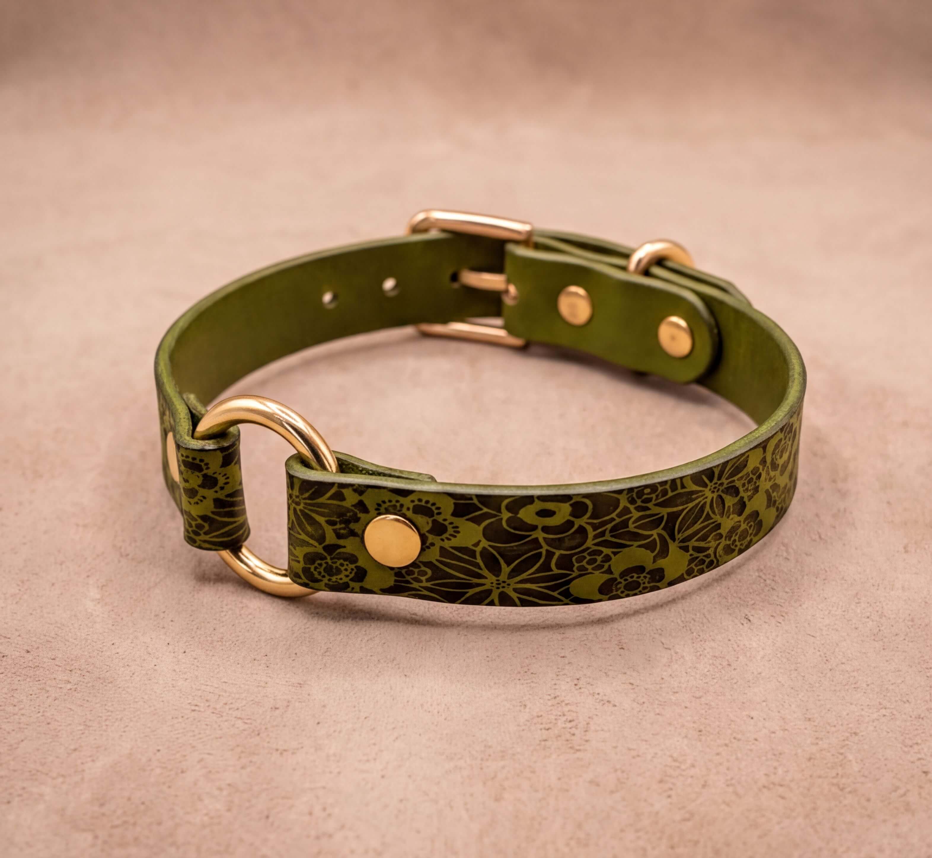 A green leather collar with a floral pattern and gold hardware, including a buckle and two rings, is displayed on a beige background.