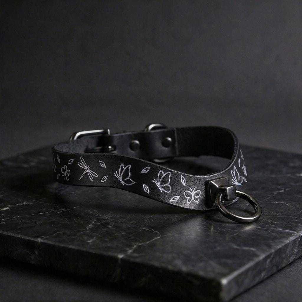 A black leather collar with a silver metal ring and a floral design on it, resting on a dark surface.