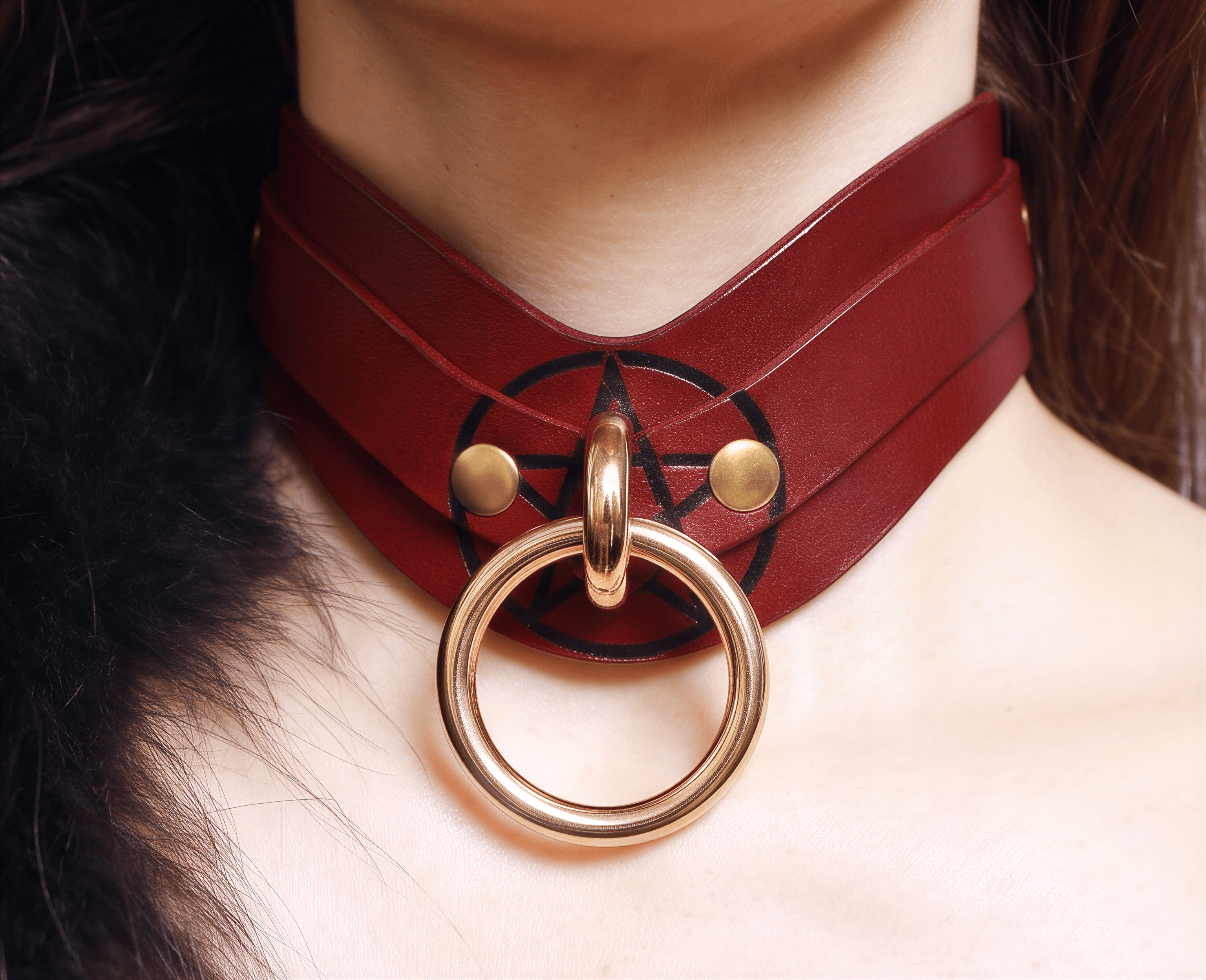 A close-up of a person's neck wearing a red leather choker necklace with a gold ring and a pentagram symbol.