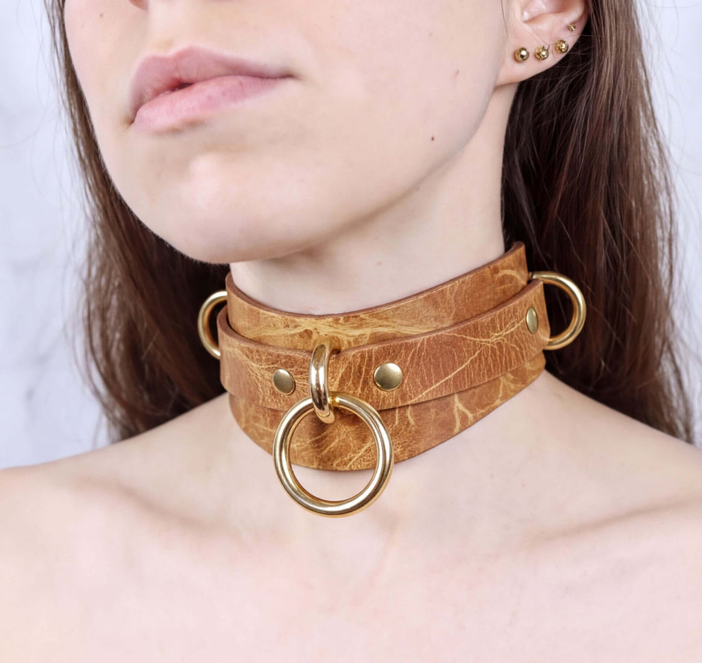 A close-up of a woman's neck and chest area wearing a brown leather choker necklace with gold-colored rings.