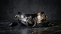 A black leather collar with silver floral designs and metal rings, placed on a dark surface with black stones or stones around it.