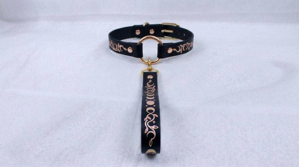 A black leather collar with a gold-colored metal clasp and a decorative metal tag attached to it, resting on a white surface.