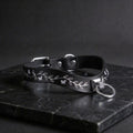 A black leather collar with a silver metal ring and leaf design on a dark surface.