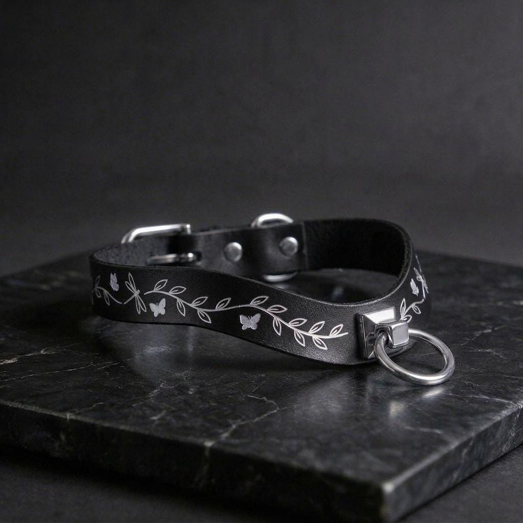 A black leather collar with a silver metal ring and leaf design on a dark surface.