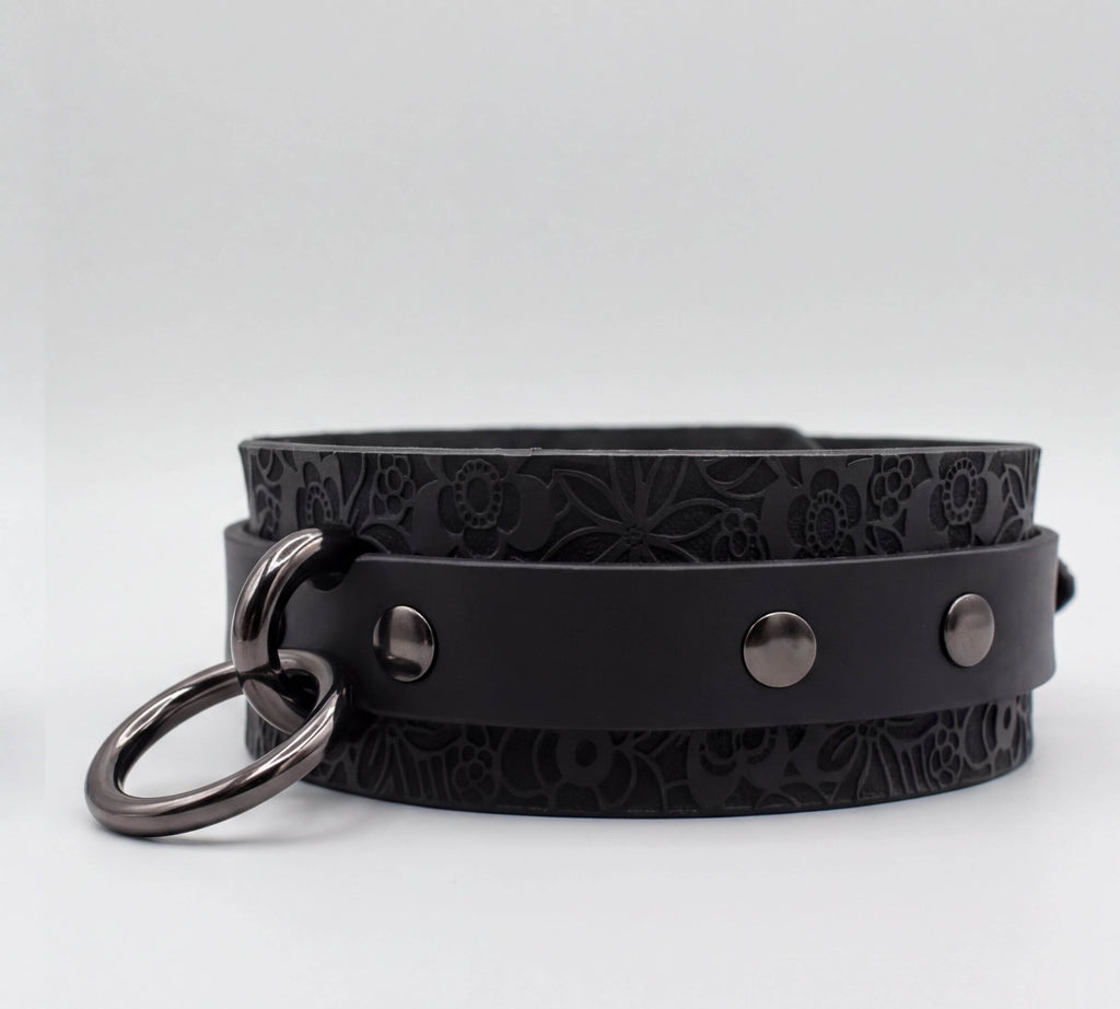 A black leather choker collar with metal rings and three circular buttons.