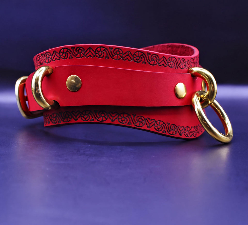 A red leather choker with gold metal rings and a decorative pattern on the edges is displayed against a dark blue background.