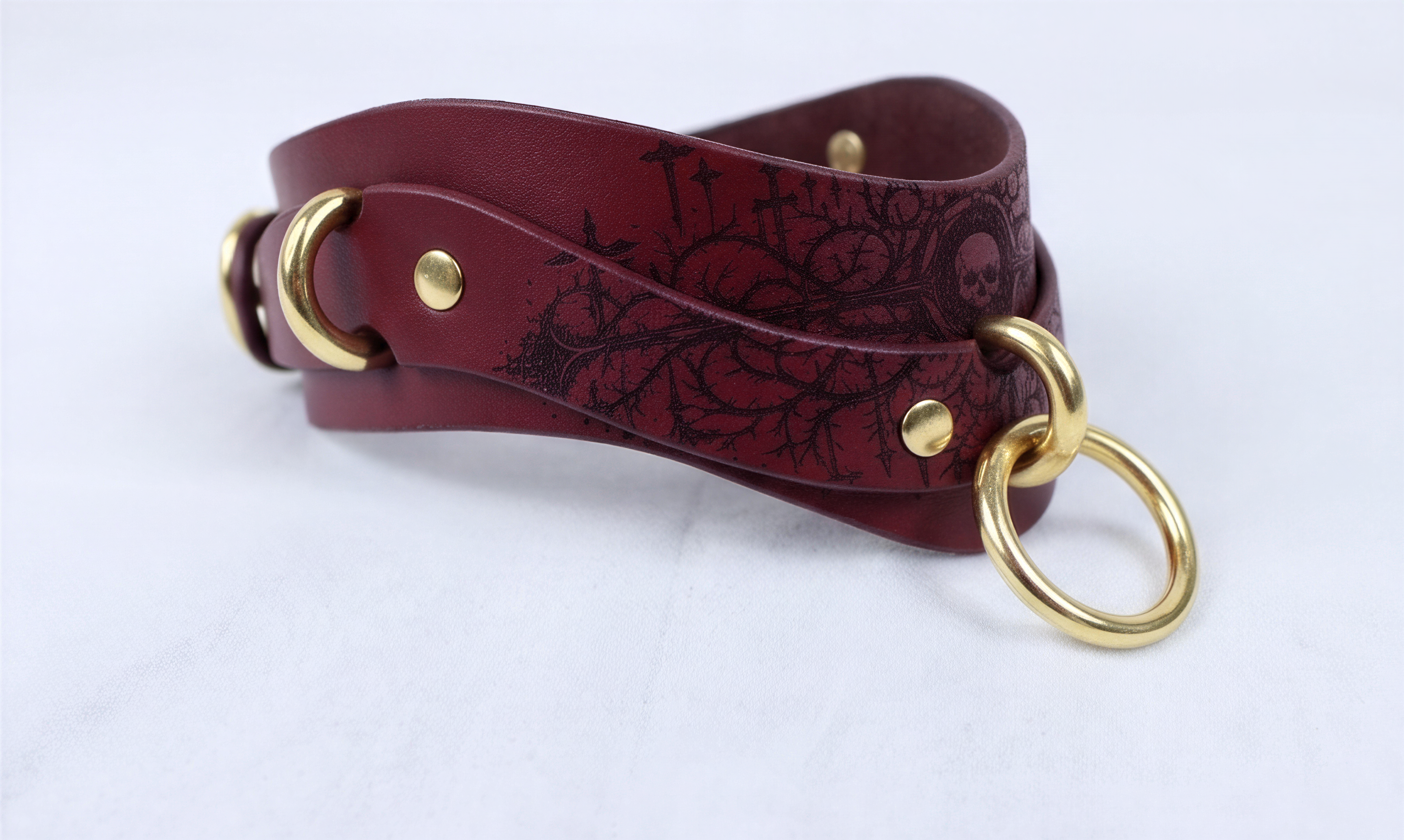 A burgundy leather choker necklace with a black floral design and gold-colored metal rings.