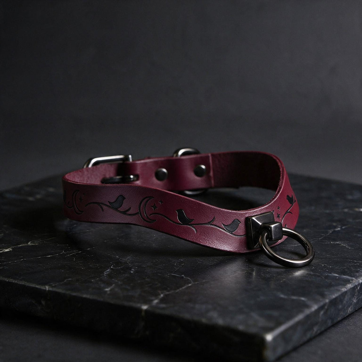 A burgundy leather collar with a metal ring and a small bird design, resting on a dark surface.