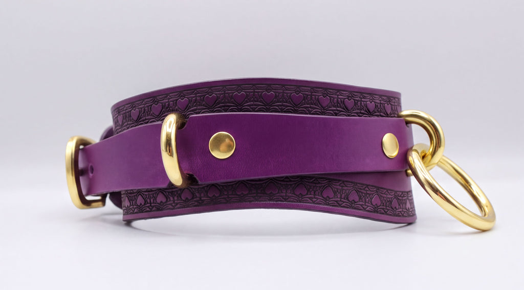 A purple leather choker collar with gold hardware, including a buckle and two rings, is shown in the image.
