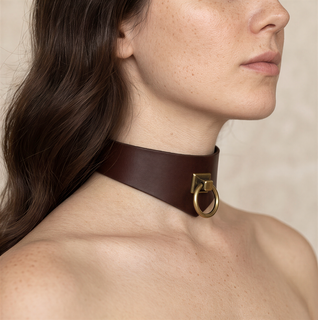 A close-up of a woman's neck and chest area wearing a brown leather choker necklace with a gold-colored metal ring.