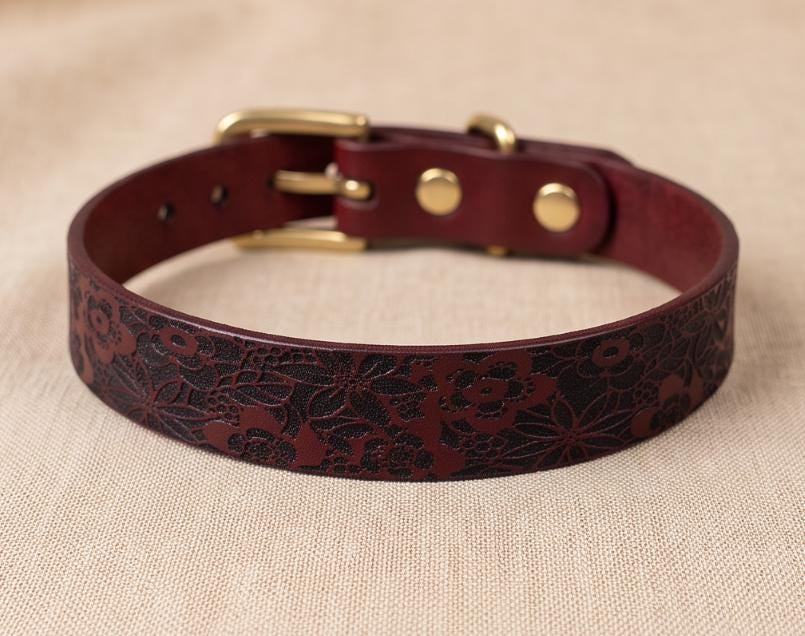 A burgundy leather dog collar with a floral pattern and a gold buckle is displayed on a beige fabric background.
