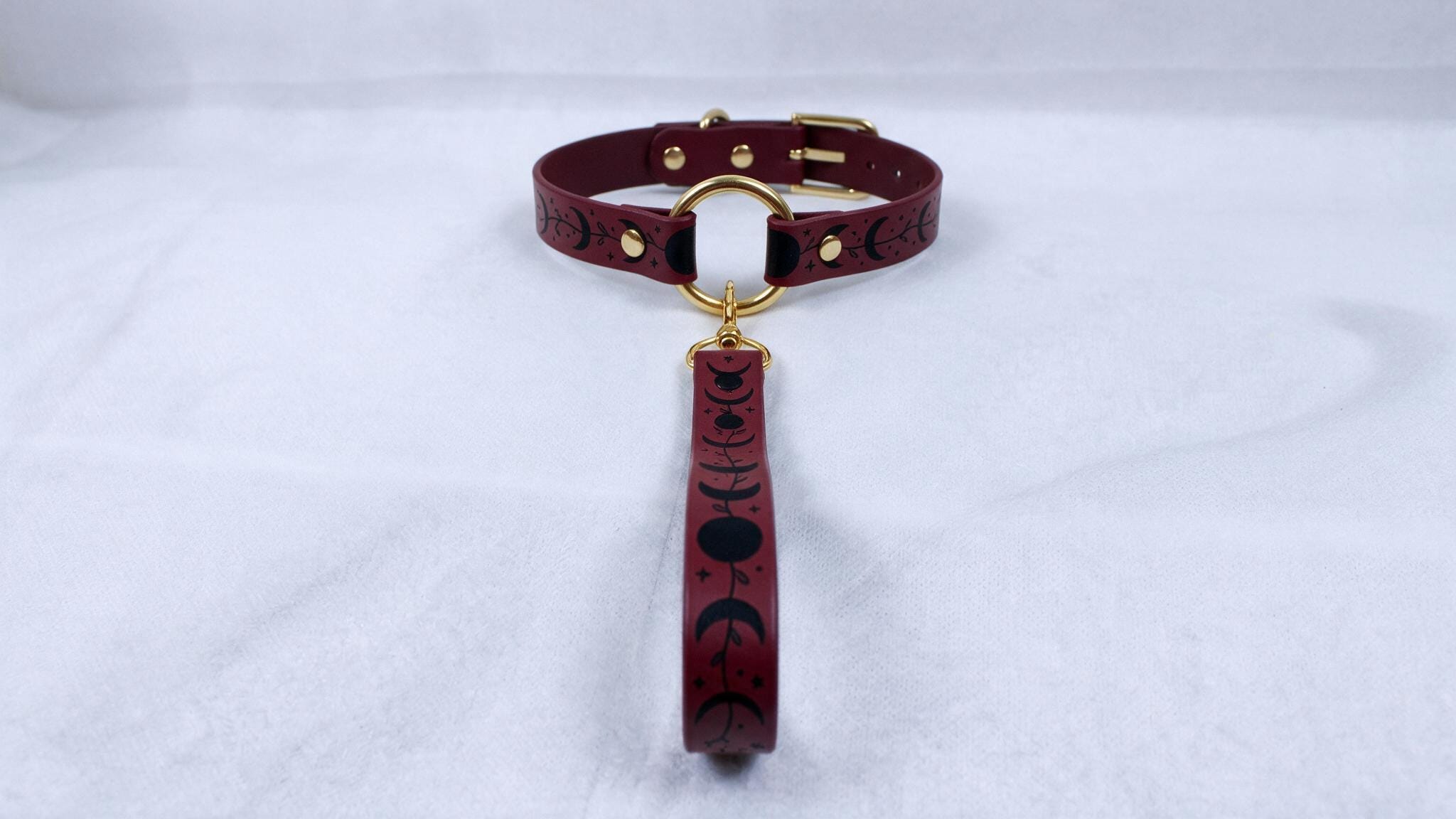 A burgundy leather collar with a gold-colored buckle and a dangling charm featuring black silhouettes of the moon and crescent moons.