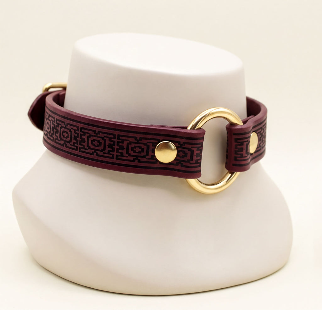 A burgundy leather choker necklace with a gold-colored circular clasp and a decorative pattern on the strap.