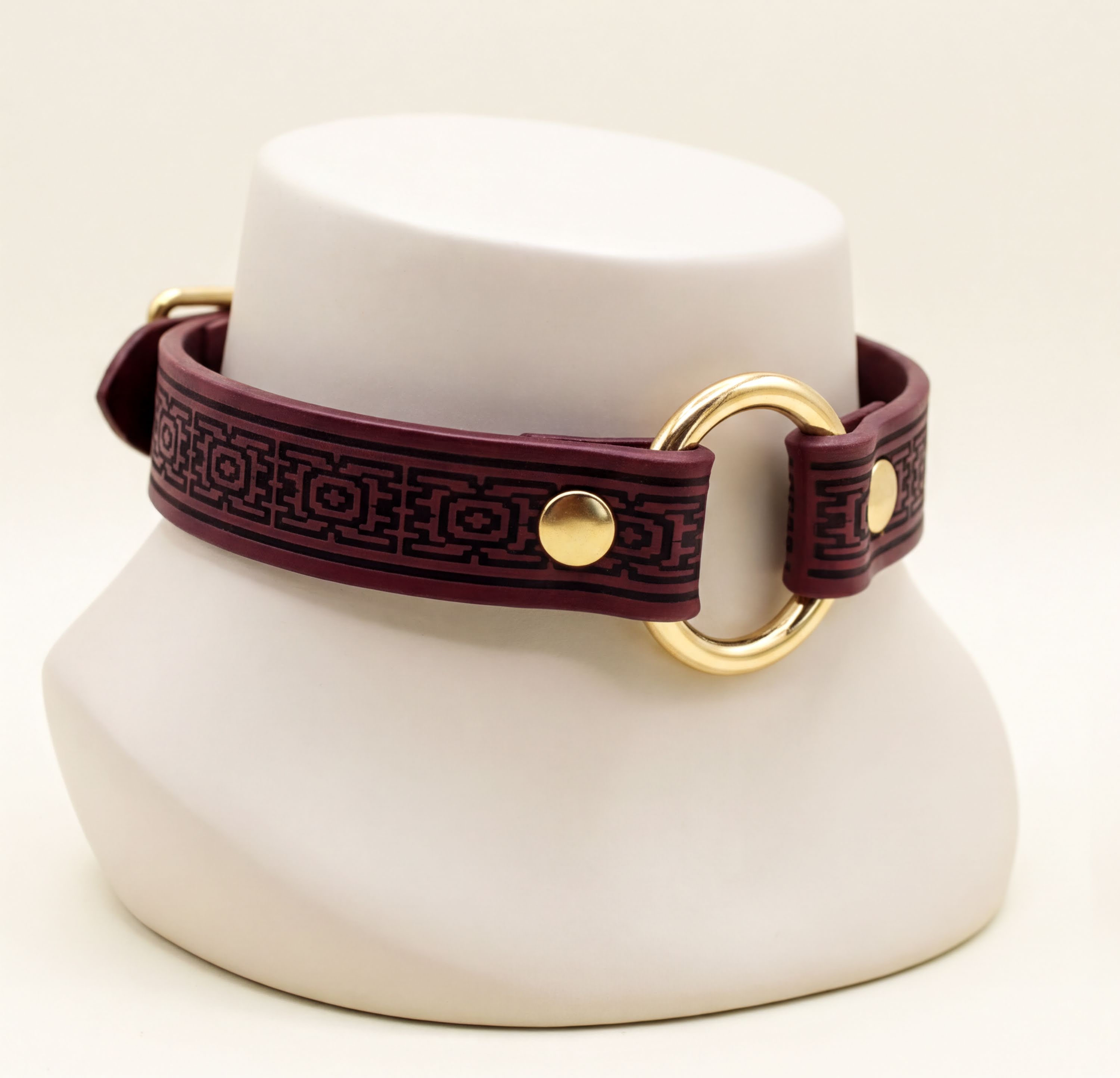 A burgundy leather choker necklace with a gold-colored circular clasp and a decorative pattern on the strap.