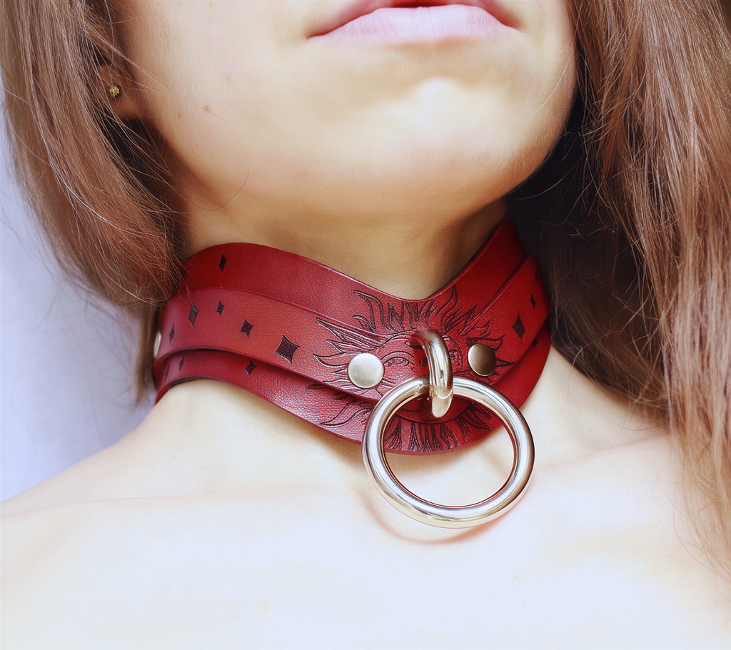 A close-up of a person's neck and chest area wearing a red leather choker necklace with a large metal ring.