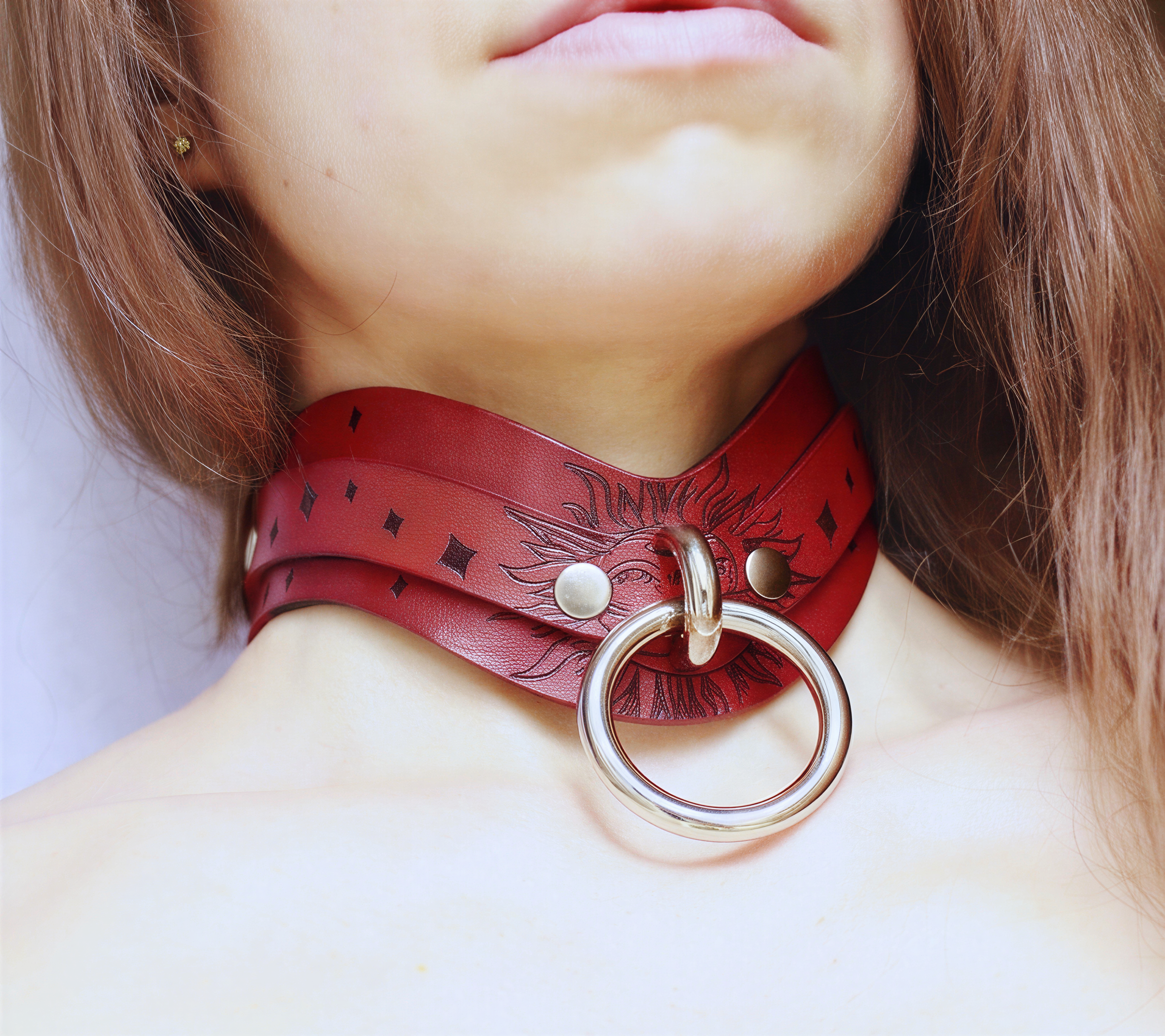 A close-up of a person's neck and chest area wearing a red leather choker necklace with a large metal ring.