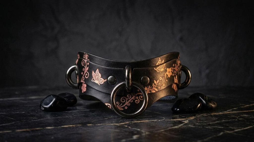 A black leather choker necklace with a floral pattern and metal rings is displayed on a dark surface, accompanied by black stones.