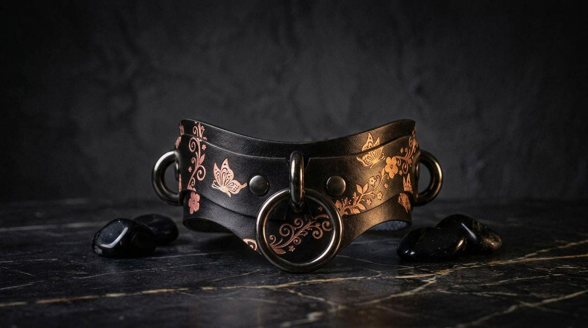 A black leather choker necklace with a floral pattern and metal rings is displayed on a dark surface, accompanied by black stones.