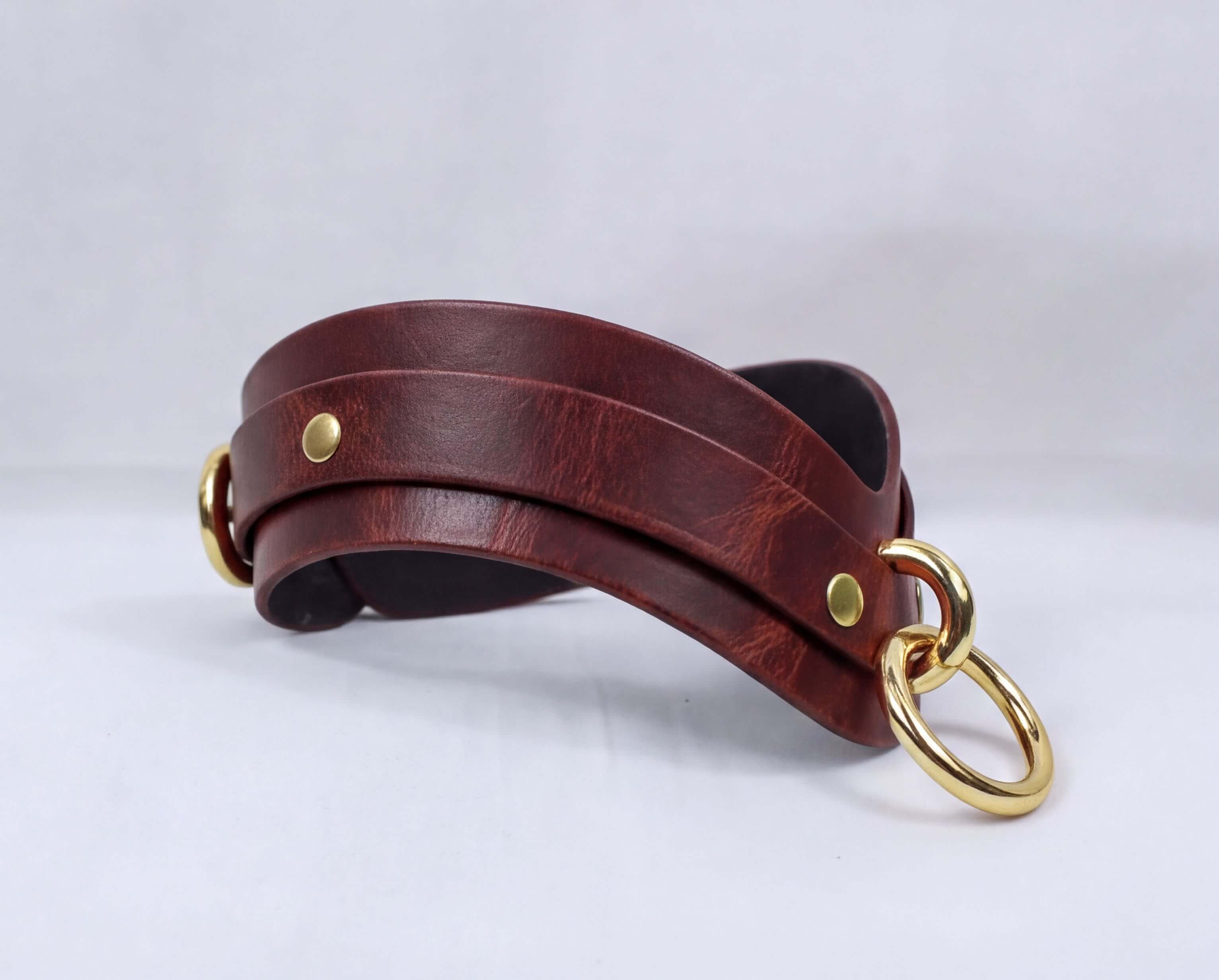 A brown leather strap with gold-colored metal rings and buckles, displayed on a white background.