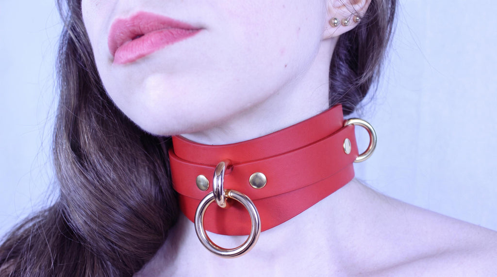 A close-up of a woman's face wearing a red leather choker necklace with gold hardware.