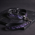 A purple leather collar with a floral pattern and a matching leash are displayed on a dark surface.
