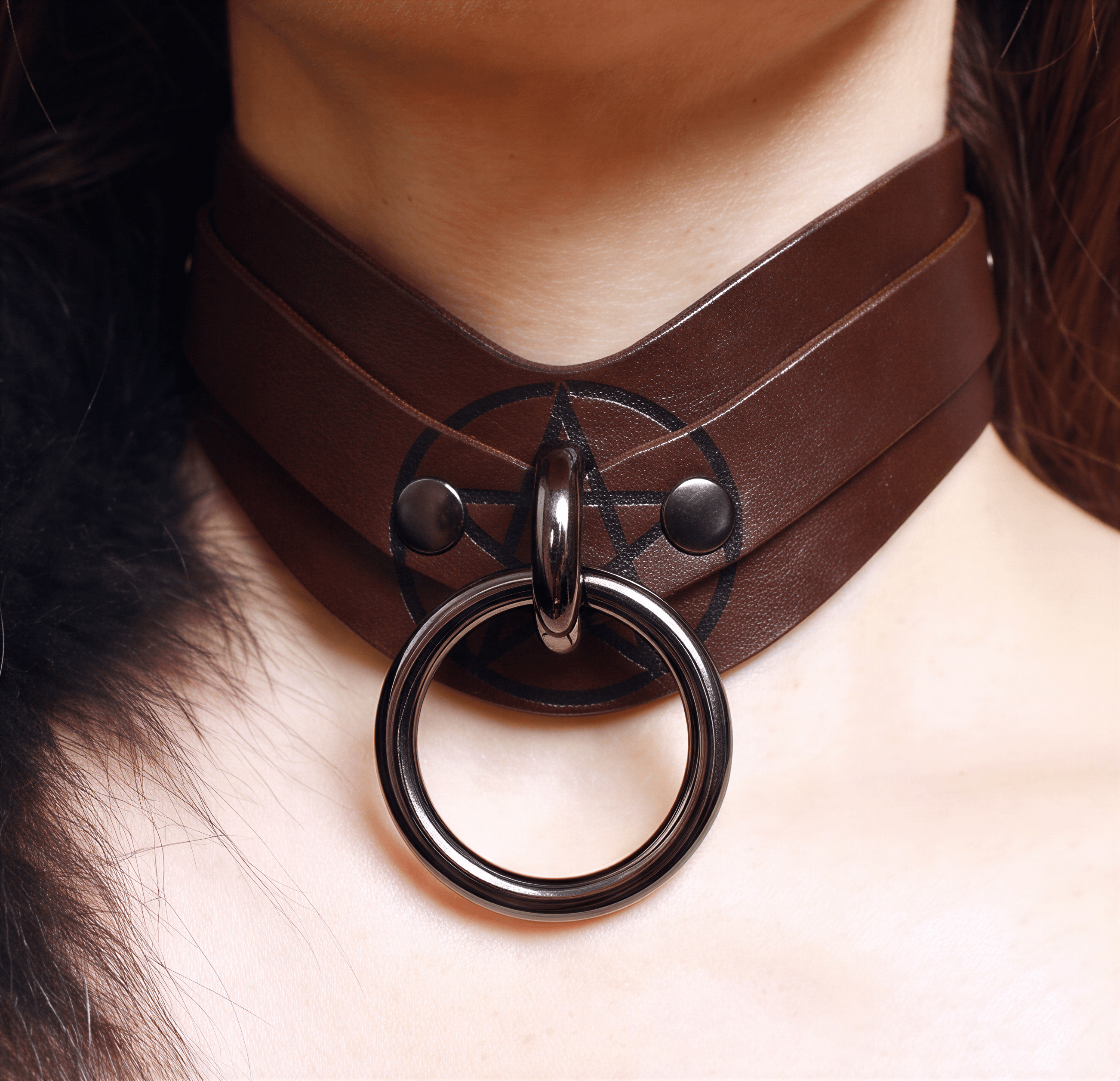 A close-up view of a person's neck and chest area, featuring a brown leather choker necklace with a metal ring and a pentagram symbol.