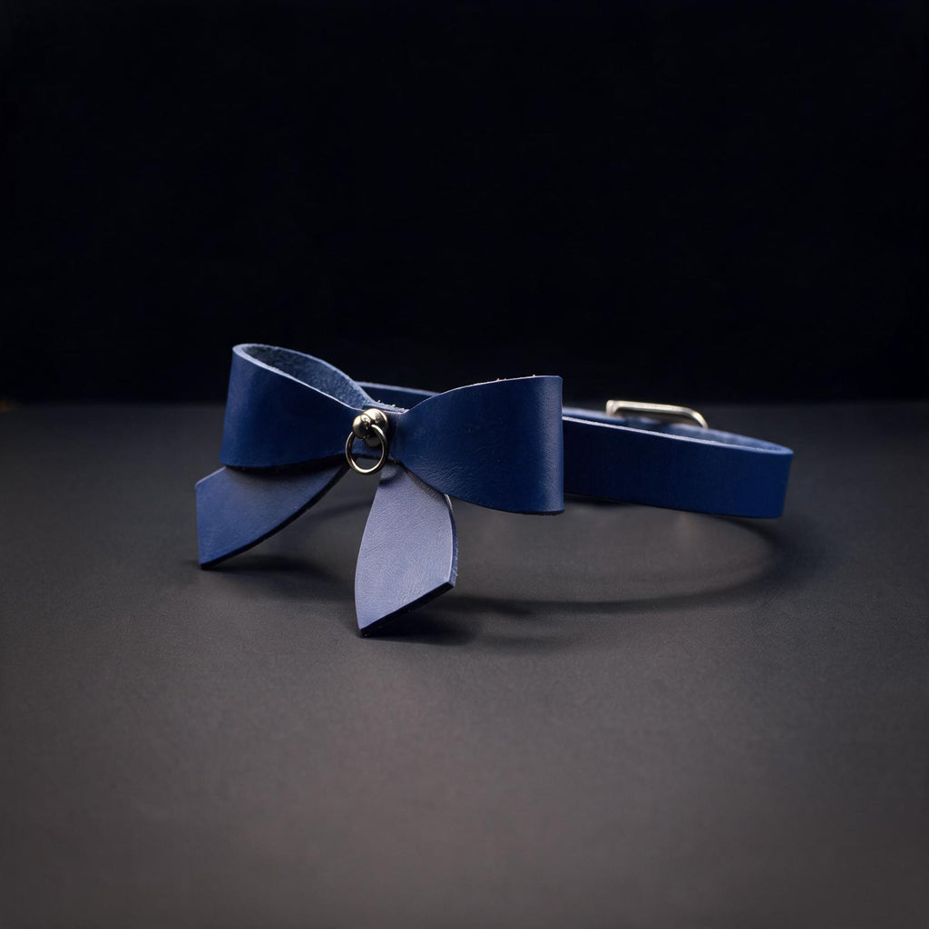 A blue leather bow tie with a metal clasp is displayed against a dark background.