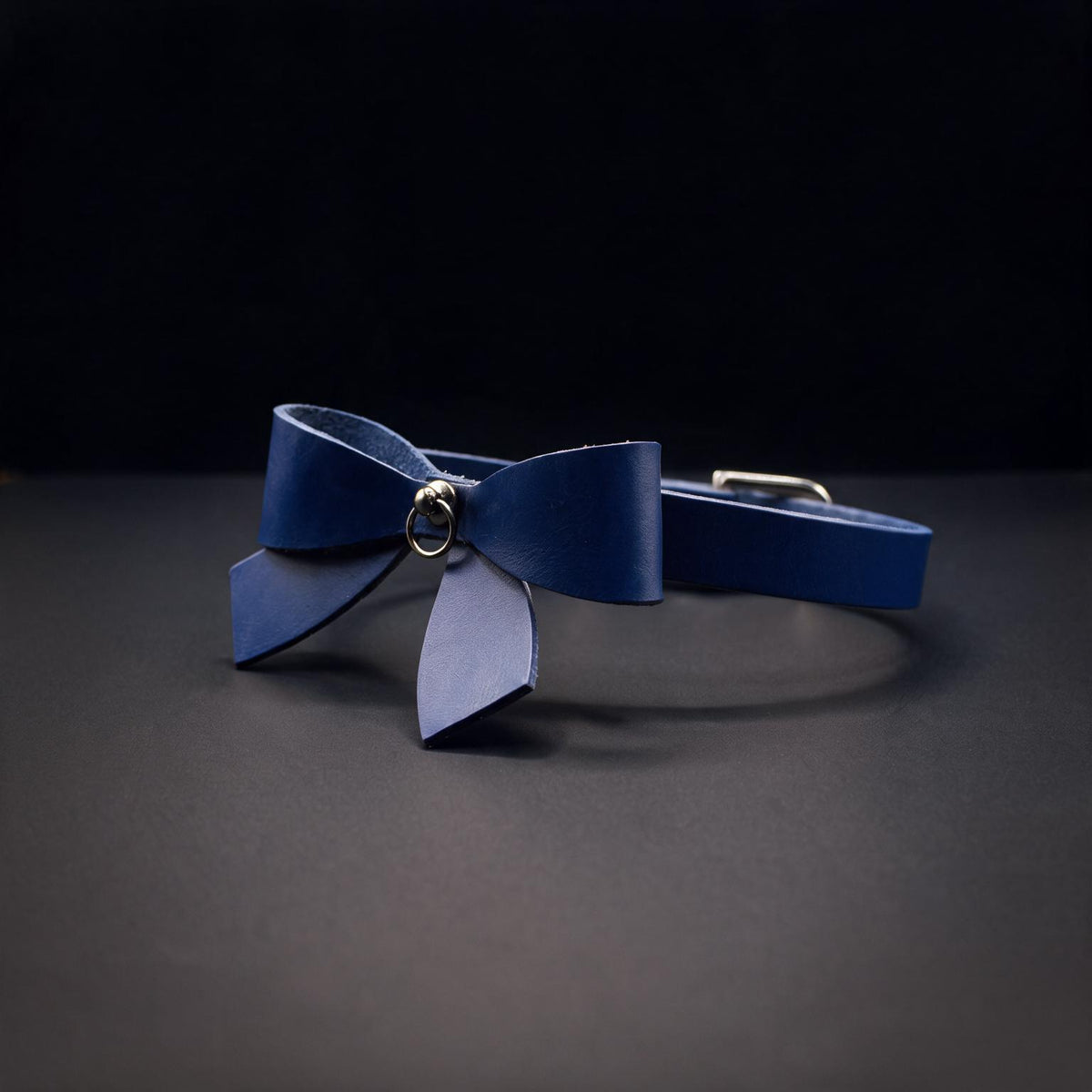 A blue leather bow tie with a metal clasp is displayed against a dark background.
