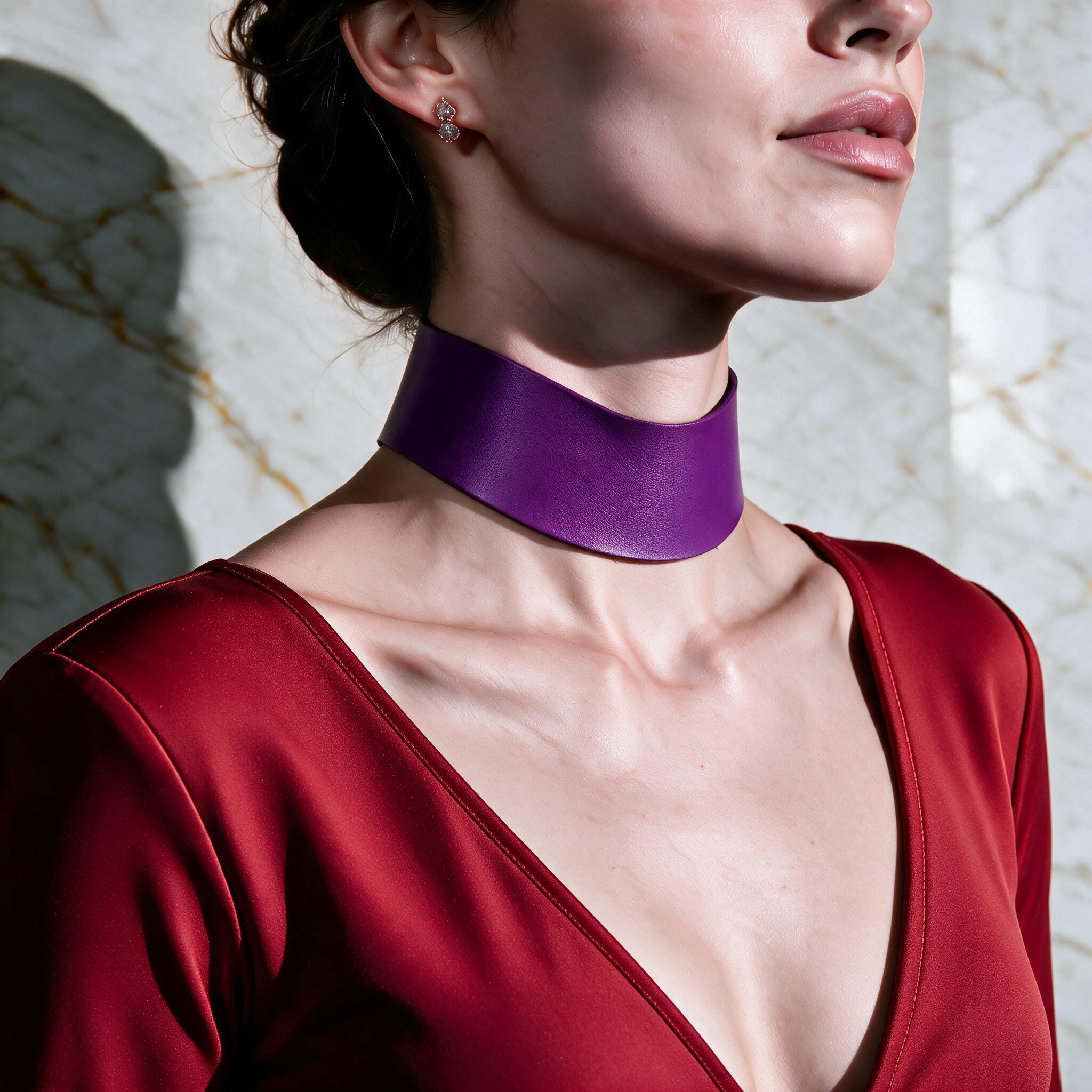 A woman wearing a red dress with a purple choker necklace stands against a marble wall.