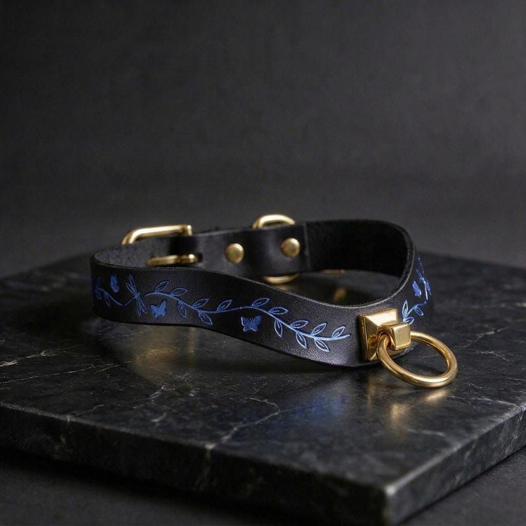 A black leather collar with a gold-colored metal ring and leaf-like designs on it, resting on a dark surface.
