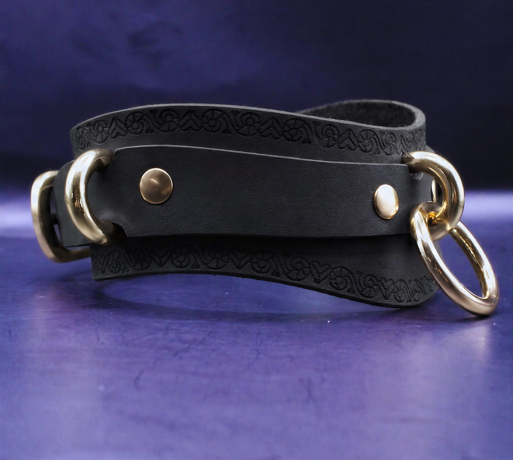 A black leather choker with gold metal rings and a decorative pattern on the strap is displayed against a purple background.