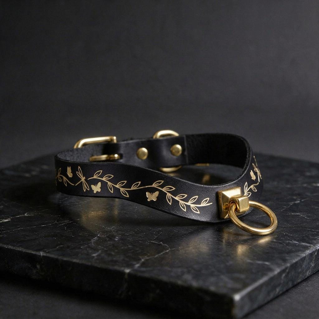 A black leather collar with gold leaf and butterfly designs, featuring a gold-colored metal ring and buckle.