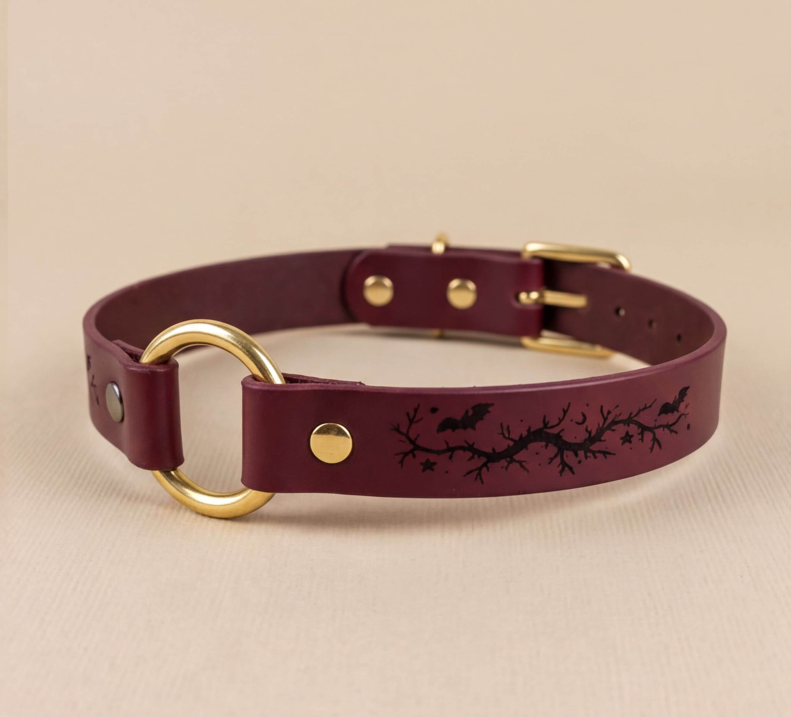 A burgundy leather dog collar with a gold ring and a design of bats and branches on the leather.