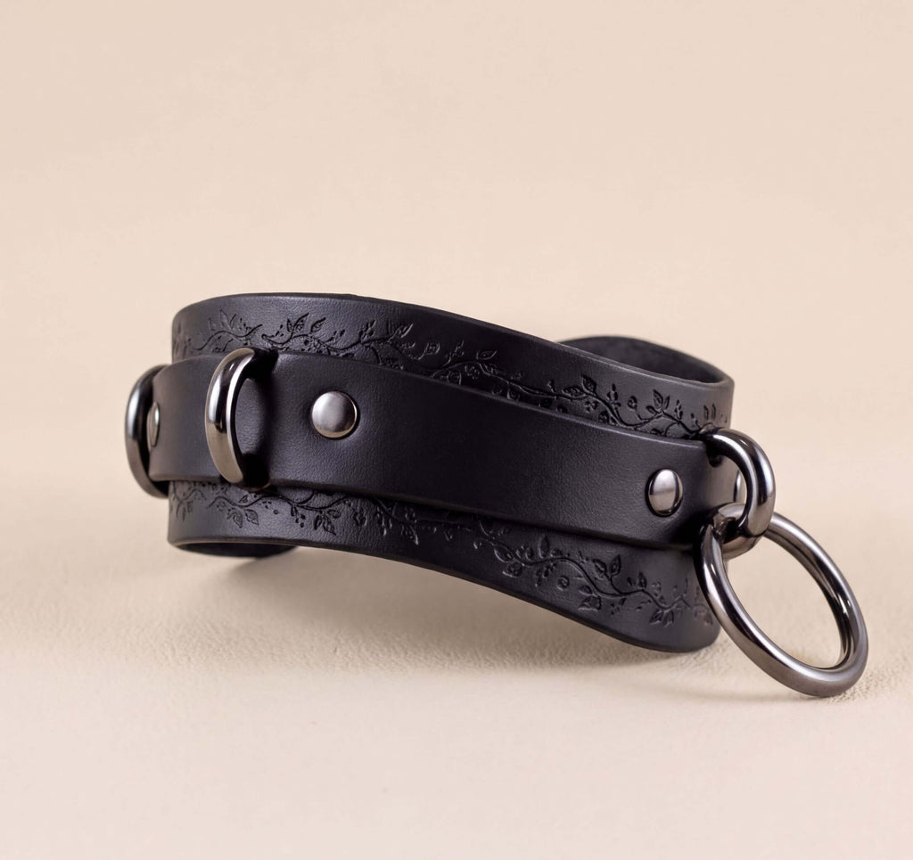 A black leather choker with metal rings and a decorative pattern on the straps.