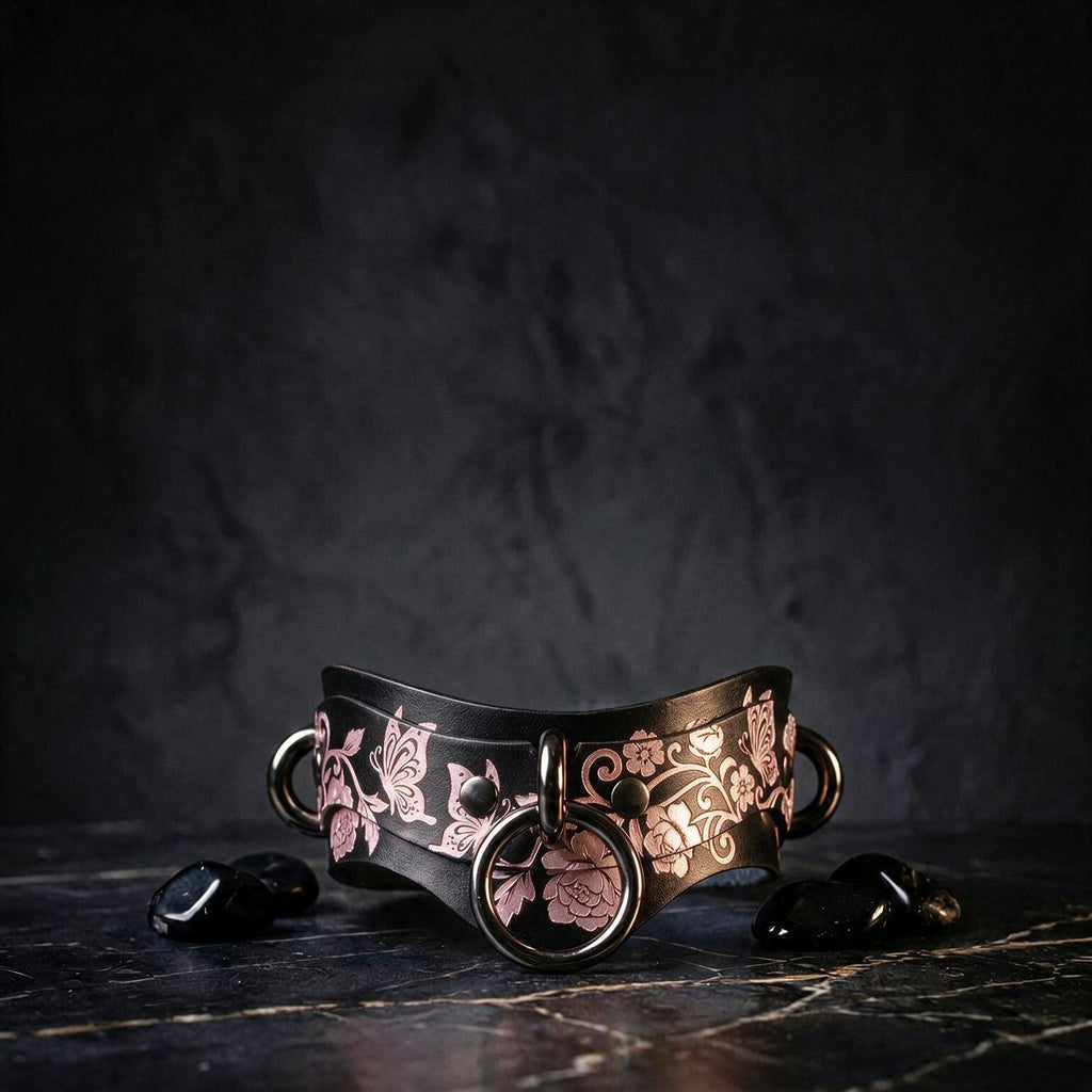 A black leather choker necklace with a floral pattern and metal rings is displayed on a dark surface, accompanied by small black stones.