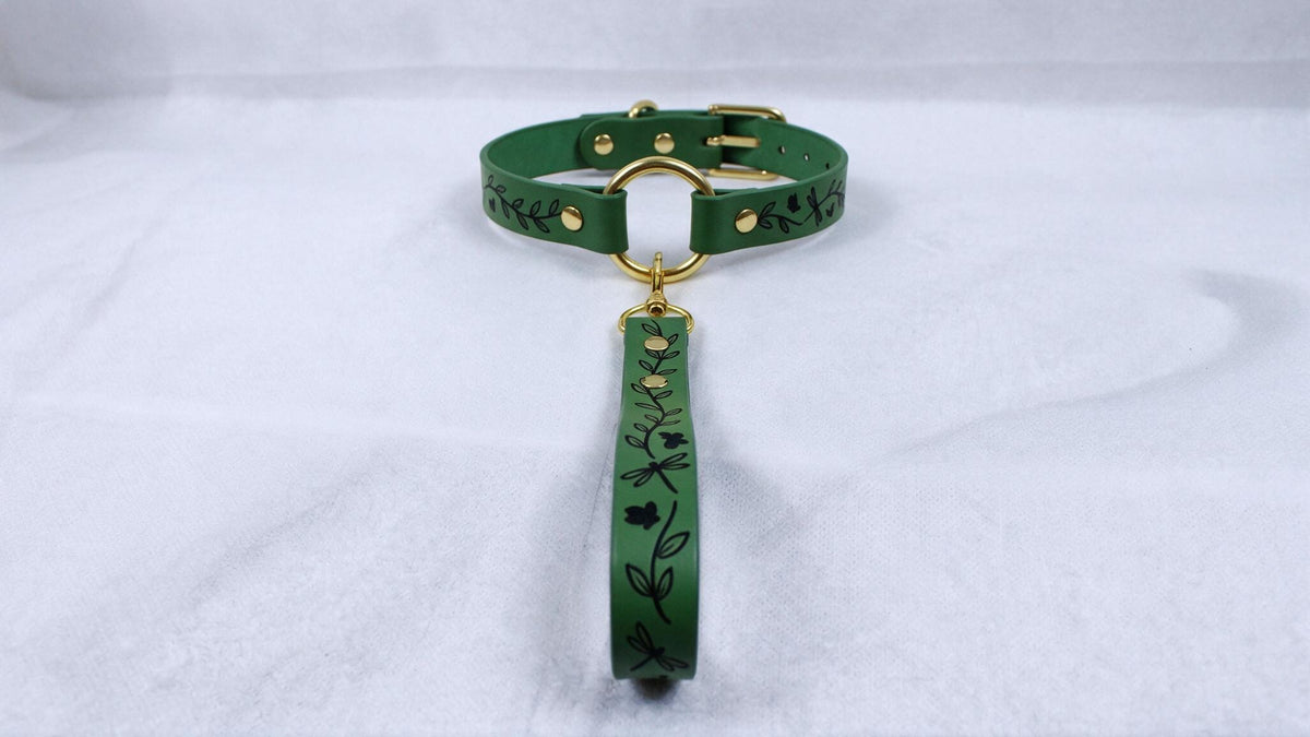 A green dog collar with a gold buckle and a black leaf design on the strap.