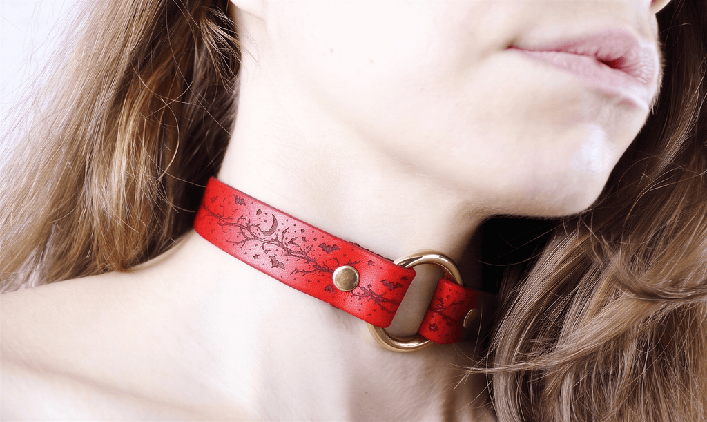 A close-up of a woman's neck and chin wearing a red choker necklace with a gold-colored metal ring.